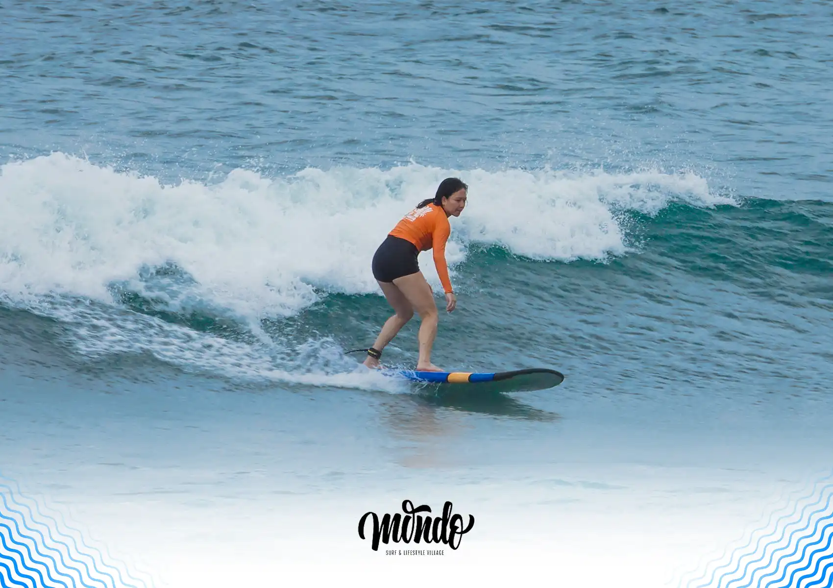 A female surfer in an orange rash guard confidently riding a gentle whitewater wave towards the shore on a blue soft-top surfboard during a lesson at a surf camp bali.