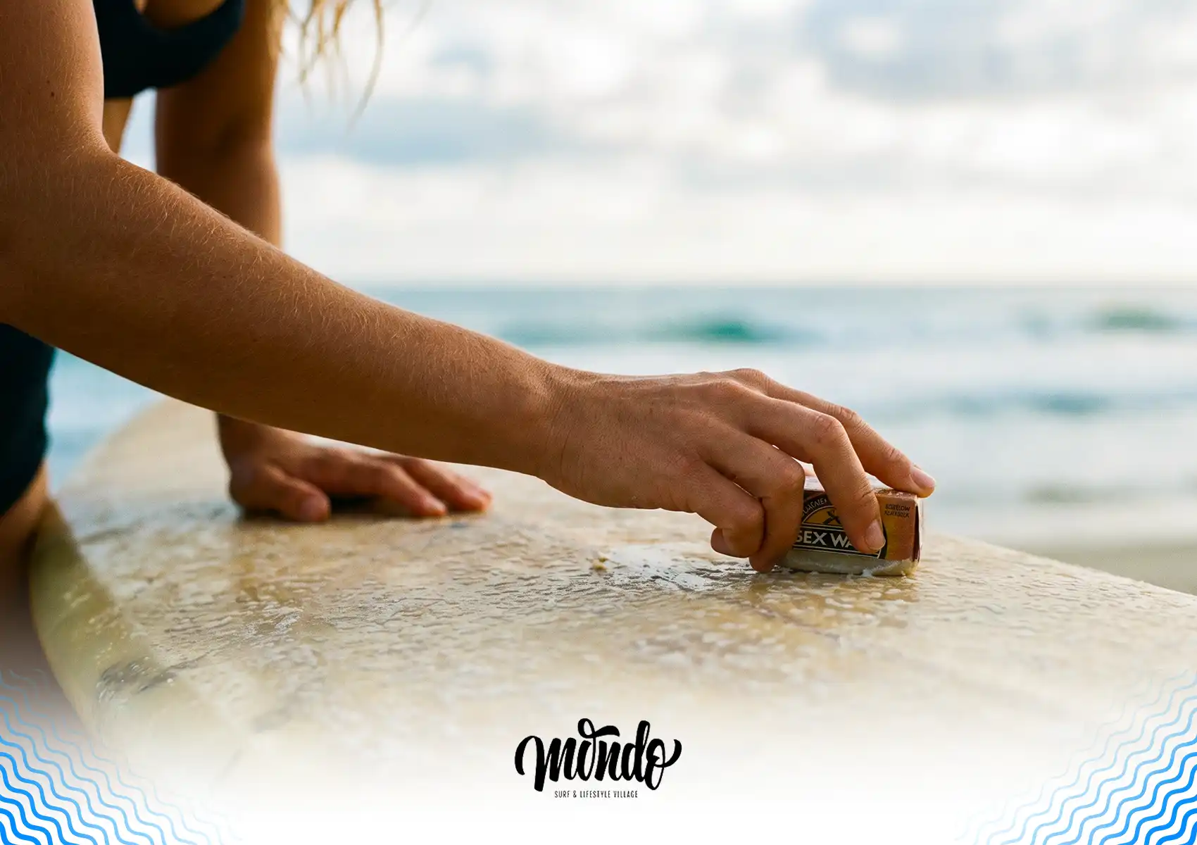A close-up of a surfer's hand applying wax to the deck of a surfboard by the ocean, preparing for an upcoming session at a surf camp bali.